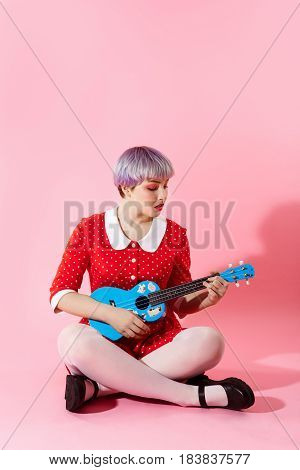Picture of beautiful dollish girl with short light violet hair wearing red dress playing blue ukulele over pink background. Copy space.