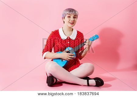 Picture of beautiful dollish girl with short light violet hair wearing red dress playing blue ukulele over pink background. Copy space.
