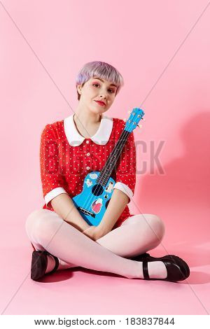Picture of beautiful dollish girl with short light violet hair wearing red dress holding blue ukulele over pink background. Copy space.