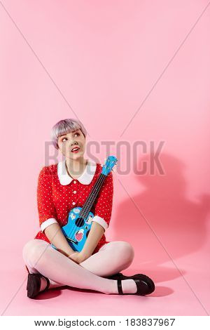 Picture of beautiful dollish girl with short light violet hair wearing red dress holding blue ukulele over pink background. Copy space.