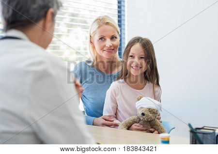 Mother and smiling daughter in a doctor office. Smiling young girl holding teddy bear toy with mother at pediatrician in hospital. Young mother and child meeting pediatrician in medical clinic.
