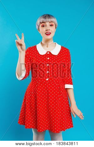 Close up portrait beautiful dollish girl with short light violet hair wearing red dress showing victory gesture over blue background. Copy space.