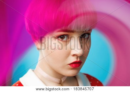 Close up artistic conceptual portrait of beautiful dollish girl with short light violet hair wearing red dress over blue background. Blurry forefront. Copy space.