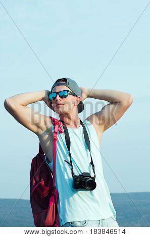 Young man caucasian traveler or photographer with muscular hands triceps biceps in stylish sunglasses and cap camera red backpack enjoy on sunny day on blue sky. Leisure and summer vacation