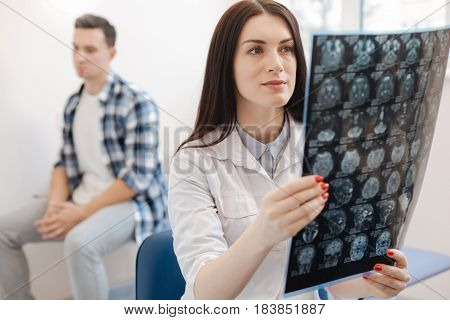 Visit to the neurologist. Beautiful attractive pleasant doctor holding an X ray photo and looking at while putting a diagnosis