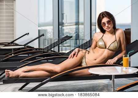 Young attractive slim girl in bikini relaxing on deck chair in wellness spa hotel resort. Near the table is a glass of orange juice