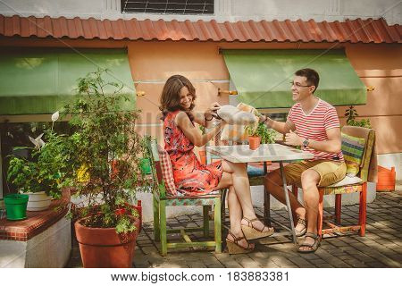 Young Funny Happy Loving Couple Sitting At Street Open-air Cafe And Fighting With Pillows Outdoors.