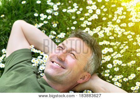 Handsome happy smiling middle-aged man lying on summer meadow green grass with daisy