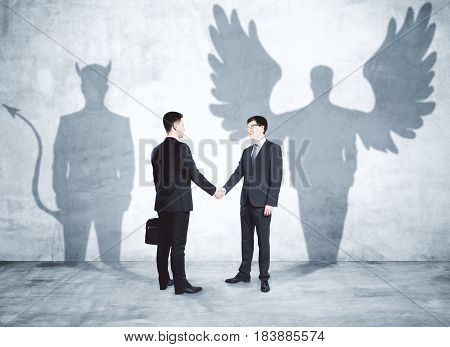 Businessmen with angel and demon shadows shaking hands in concrete interior. Partnership concept