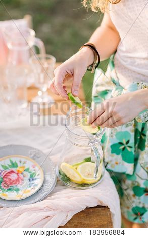 picnic, people, food, summer, holiday concept - cocktail lemonade non-alcoholic, hands of young woman mix slices of lemon, lime and mint leaves in a glass flagon, table with festive dishes, glasses.