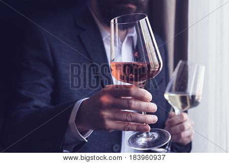 horizontal close up of a Caucasian man with beard black suit and white shirt offering a tall glass of rose wine at an event by the window natural light