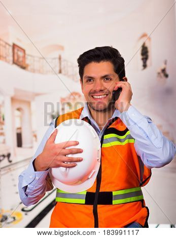 Smiling engineer with a helmet in his hand at construction site while he is using his celllphone.