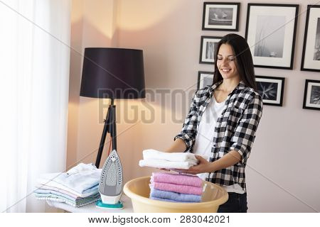 Beautiful Young Woman Standing Next To An Ironing Board, Folding Clean, Ironed Towels Into A Washbow