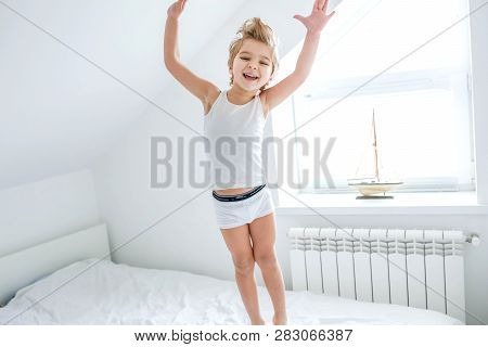 Happy Boy Playing In White Bedroom. Little Boy Brother Play On The Bed Wearing Pajamas. Family At Ho