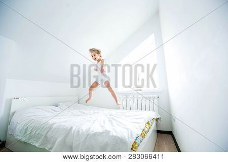 Happy Boy Playing In White Bedroom. Little Boy Brother Play On The Bed Wearing Pajamas. Family At Ho