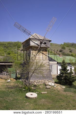 Close-up Of An Old Wooden Mill With Blades. In The Foreground Are Round Millstones On Green Grass, G