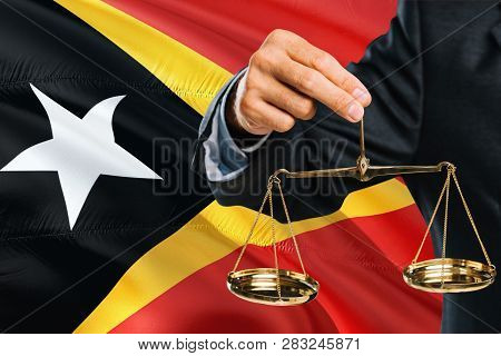 Timorese Judge Is Holding Golden Scales Of Justice With East Timor Waving Flag Background. Equality 