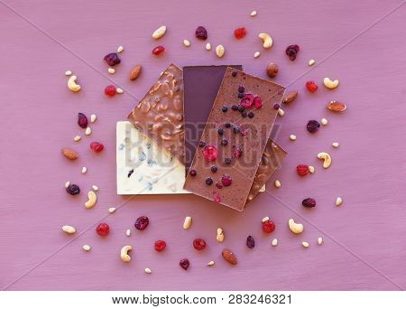 Chocolate On A Violet Background, Surrounded By Nuts And Dried Fruits. Chocolate. Chocolate Bar. Nut