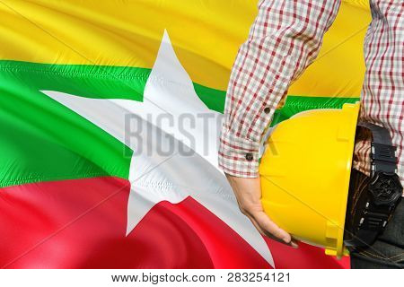 Burmese Engineer Is Holding Yellow Safety Helmet With Waving Myanmar Flag Background. Construction A