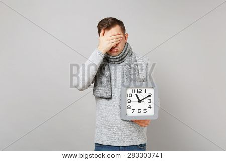 Tired Young Man In Gray Sweater, Scarf Covering Face With Hand Hold Square Clock Isolated On Grey Ba