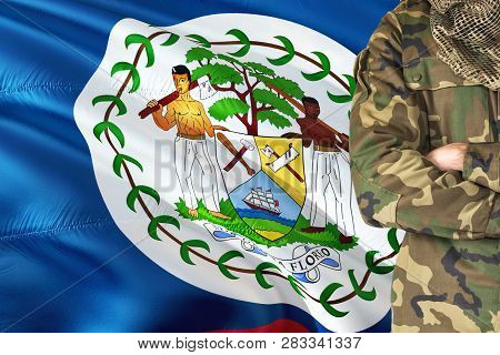Crossed Arms Belizean Soldier With National Waving Flag On Background - Belize Military Theme.