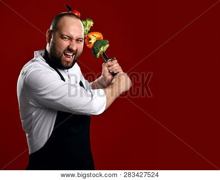 Bearded Professional Chef In White Skirt And Apron Going To Fight With Skewer With Vegetables On It 