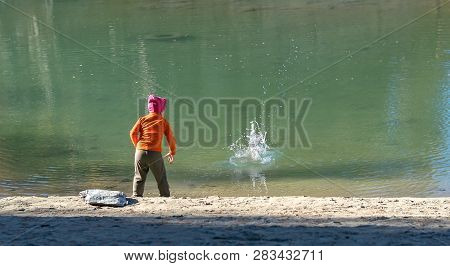 A Little Girl, A Child, In Warm Clothes And A Red Cap Throws Stones Into The Water From The Sandy Sh