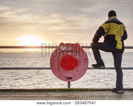 Man In Trekking Suit In Harbor In Purple Daybreak. Touristic Mole Wet Wooden Floor Above Sea.