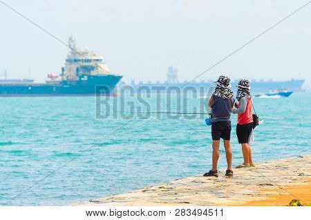 Fishermen Fishing From The Embankment Of Singapore Harbor. Commercial Cargo Ships In The Background