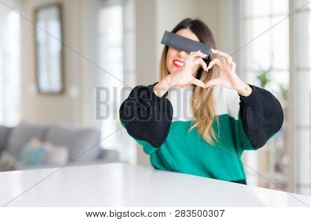 Young beautiful woman blindfold eyes with black censor band at home smiling in love showing heart symbol and shape with hands. Romantic concept.