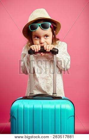 Bored Little Girl With Blue Suitcase, Wears A Straw Hat And Sunglasses, Expresses Puzzlement And Con