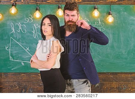 Man With Beard And Sexy Girl Teacher Stand In Classroom, Chalkboard On Background. Teacher And Stric