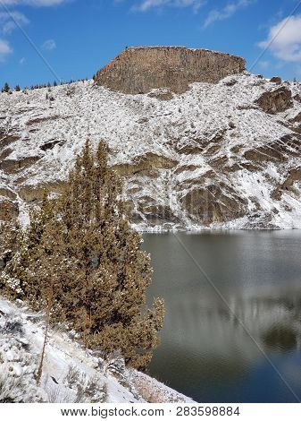 A Lone Juniper Tree In The Foreground Of An Basalt Outcropping On A Hill With A Fresh Layer Of Snow 
