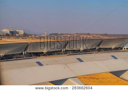 Aircraft Wing With Breaking Flaps Extended During Landing With Airport Buildings And Blue Sky In Bac