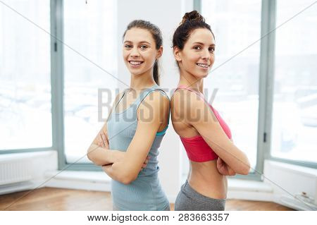 Waist Up Portrait Of Two Fit Young Women Standing Back To Back And Looking At Camera After Workout I