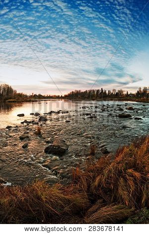 The Autumn Sun Sets Behind The River And The Light Reflects On The Water. The Rapids Create Whirly W