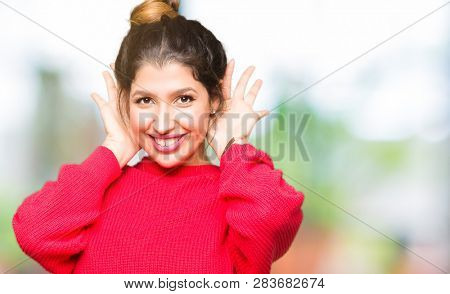 Young beautiful woman wearing red sweater and bun Trying to hear both hands on ear gesture, curious for gossip. Hearing problem, deaf