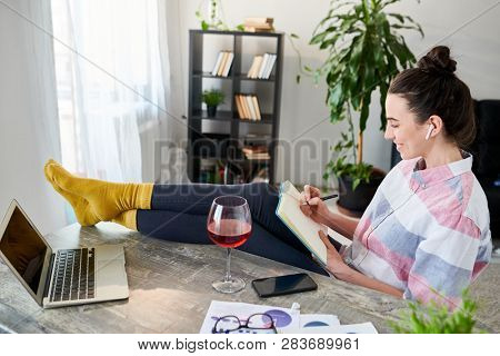 High angle portrait of relaxed young woman writing in journal with feet on desk, copy space