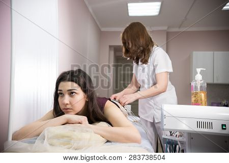 Cosmetology Beauty Clinic. Young Woman Taking Myostimulation And Doctor Preparing The Procedure At T