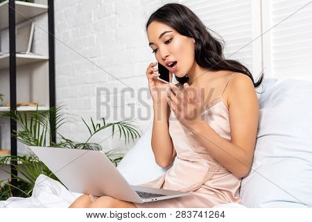 Beautiful Asian Freelancer Using Laptop And Emotionaly Talking On Smartphone While Staying In Bed