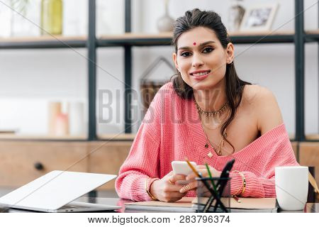 Happy Indian Student With Bindi Using Smartphone At Table With Laptop