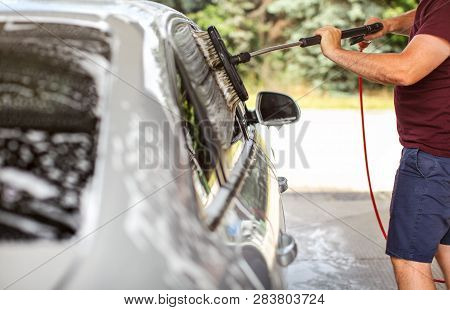 Young Man In Shorts And T Shirt Washing His Car In Self Serve Carwash, Cleaning Side Windows With Br