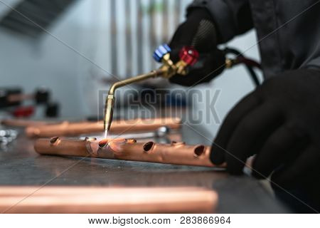 Worker Is Soldering A Pipe By A Blow Lamp On A Factory Workbench Background. Pipework.