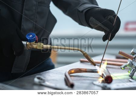 Worker Is Soldering A Pipe By A Blow Lamp On A Factory Workbench Background. Pipework.