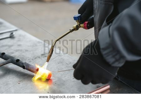 Worker Is Soldering A Pipe By A Blow Lamp On A Factory Workbench Background. Pipework.