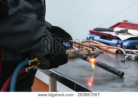 Worker Is Soldering A Pipe By A Blow Lamp On A Factory Workbench Background. Pipework.