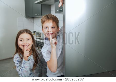 Two cute kids hiding behind the fridge in the kitchen. Childhood. Kid's fashion.