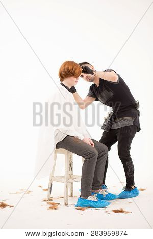 Ginger Man In A White Cape Sits At The Stool While Barber Does Hair Trimming. Full Length Of Handsom