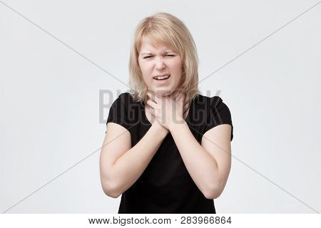 Young Blonde Woman In Black T-shirt Having Pain In Throat Clenching Hands On It.