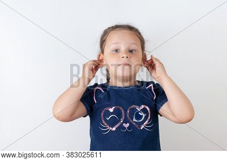A Girl In A Blue Denim Dress Holds Her Ears And Sticks Them Out. Smiling Child On A White Background